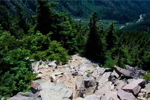 A rocky, very steep trail travels toward toward green fir trees with a winding river in the valley below