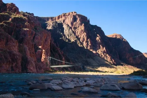 Cliffs rise above a silver suspension bridge above a shadowy river