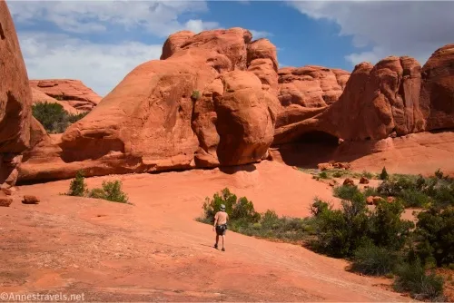 A hiker walks across slickrock toward slickrock domes and an arch with trees to his right