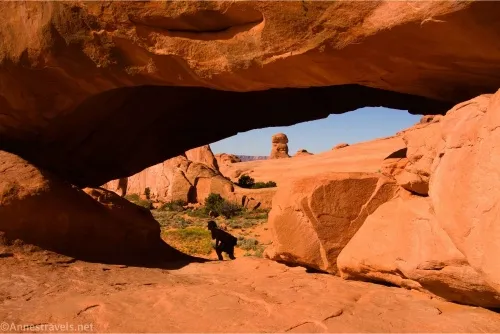 A hiker crouches under the opening of an arch with rock formations visible through the arch