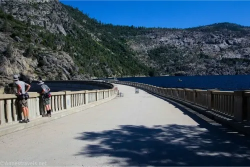 Two hikers stand along the railing of a broad top of the dam with the reservoir on the right and hills beyond