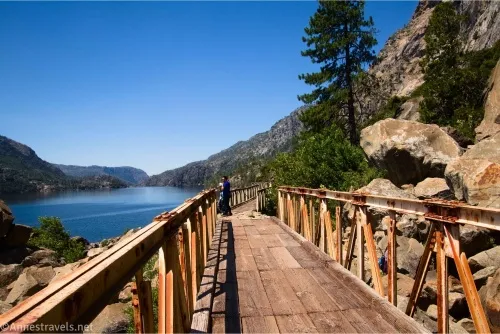 A bridge with rusty railings and a wood floor between a tree and rocks on the right and a blue reservoir on the left