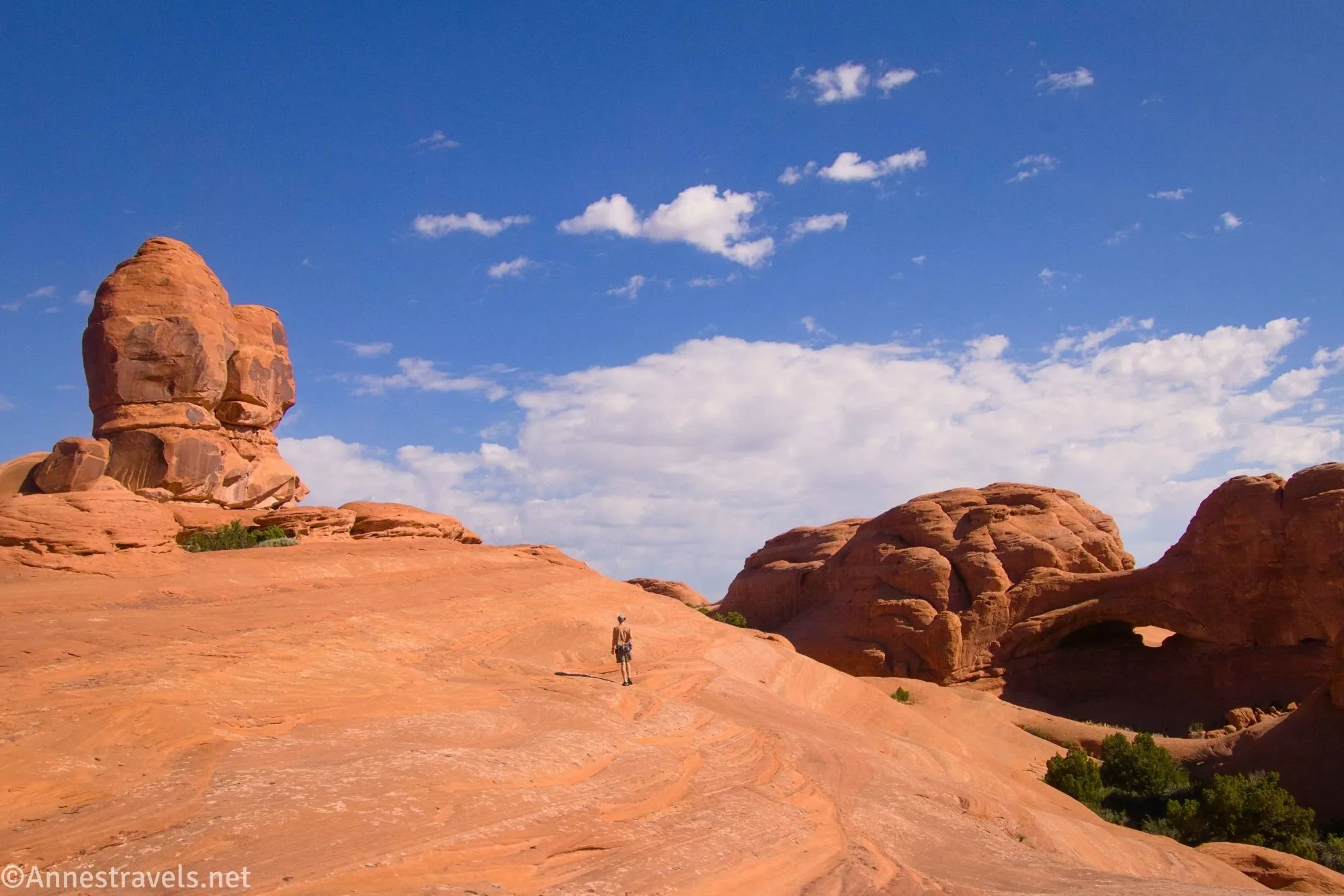A rock formation rises on the left, a hiker stands on an expanse of slickrock, and Eye of the Whale Arch sits on the right