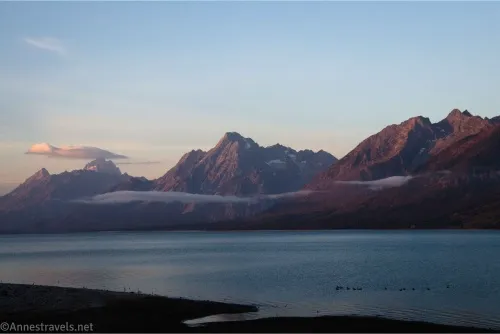 Sunrise turns craggy peaks pink while low-hanging clouds float over a peaceful lake 
