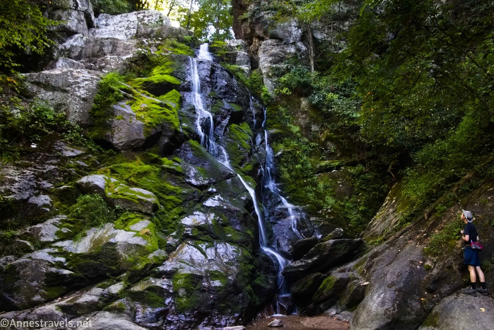 A hiker looks up at a lovely waterfall tumbling down a mossy rock face
