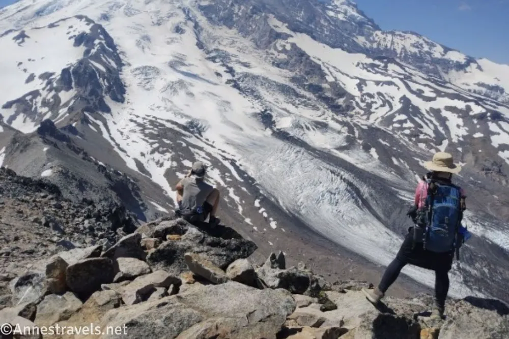 Two hikers stand on a rocky outcrop taking pictures of a glaciated volcano