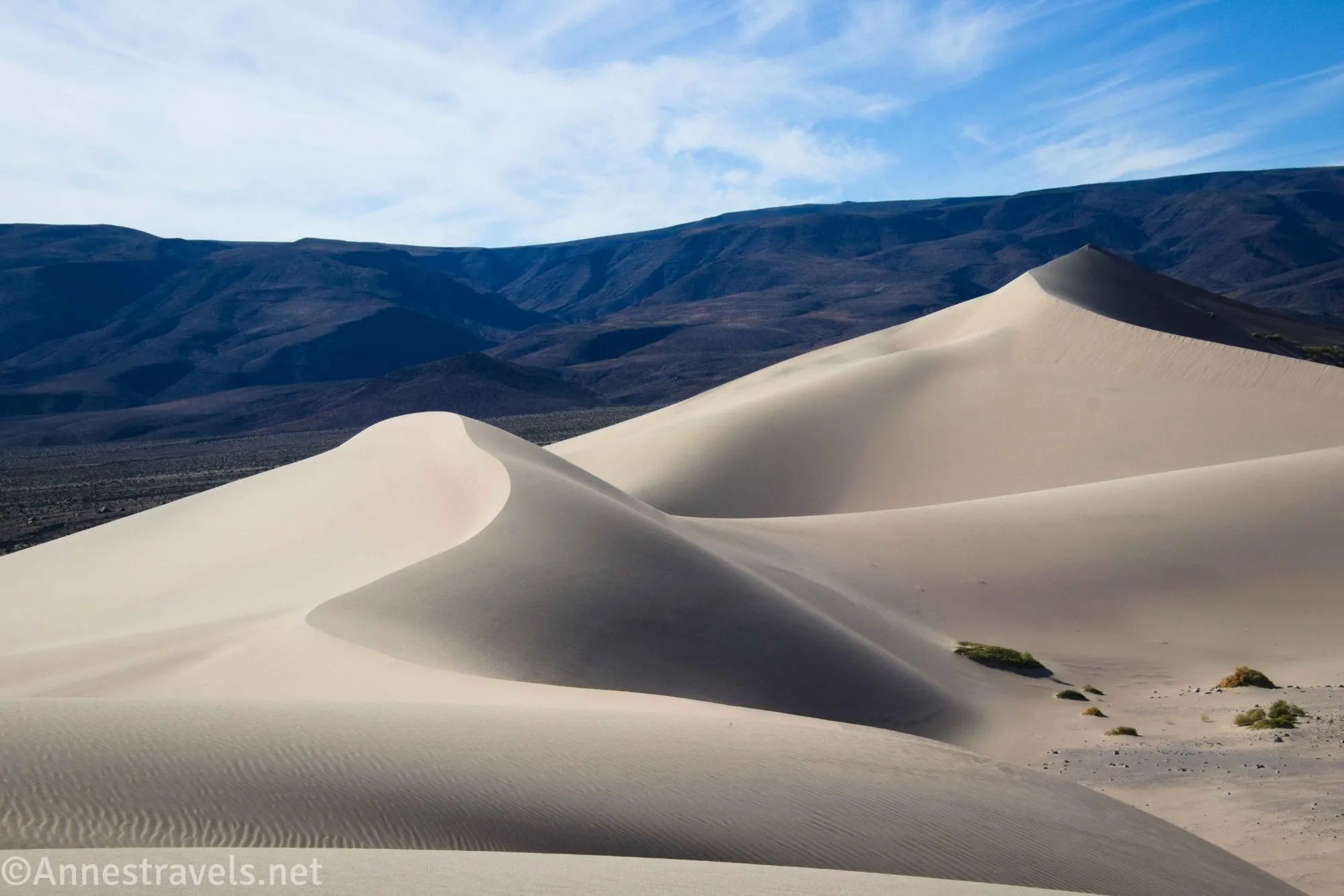 Two sand dunes below dark mountains and cloudy blue skies 