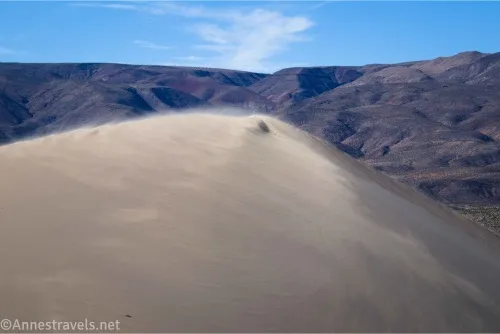 Sand blows across the top of a sand dune with mountains beyond