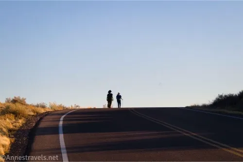 Two silhouettes walk across the top of a hill on a paved road 