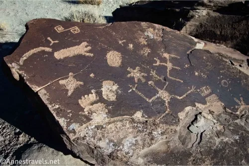 A boulder with various rock art on it, including a human, a bat, a sun, a foot, and a geometric swirl 