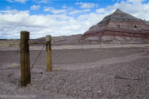 Two fenceposts hold up a wire fence with colorful striped badlands and clouds in the distance