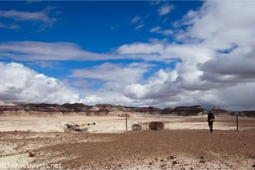 A hiker walks by a roll of fence wire and fenceposts with badlands beyond and puffy white clouds in the sky