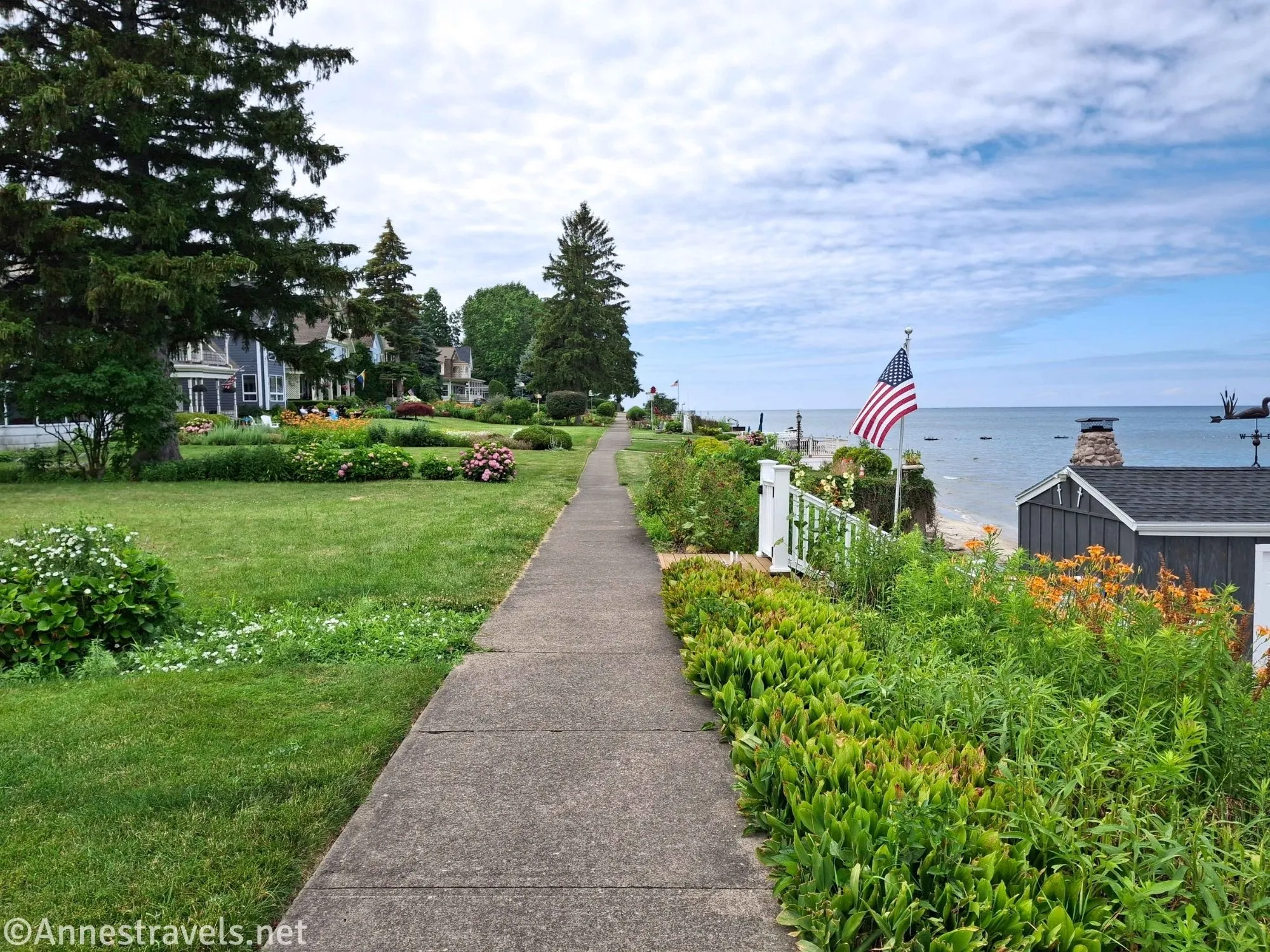 A sidewalk travels between gardens and yards beside Lake Ontario with an American Flog in one of the gardens