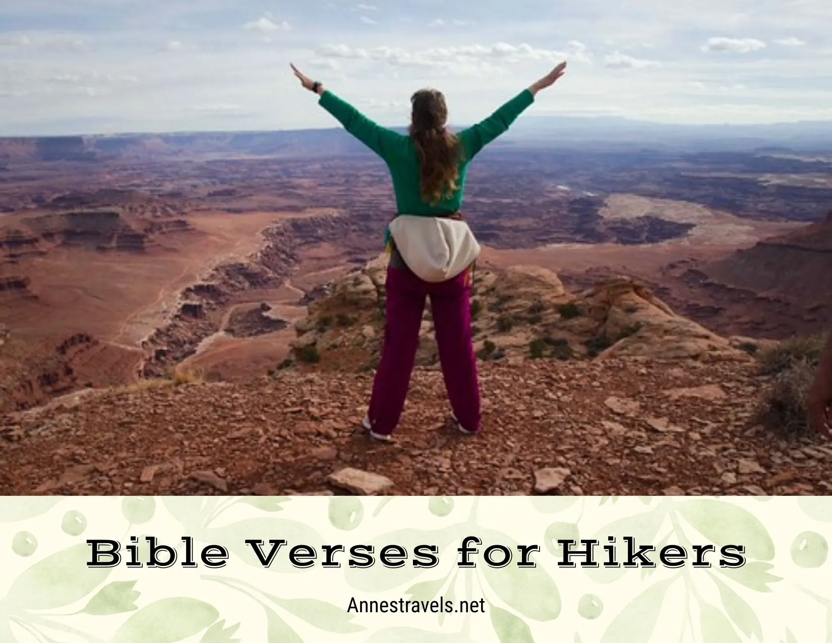A hiker with brown hair and a green shirt and pink slacks with her hands held high above a rugged canyon 