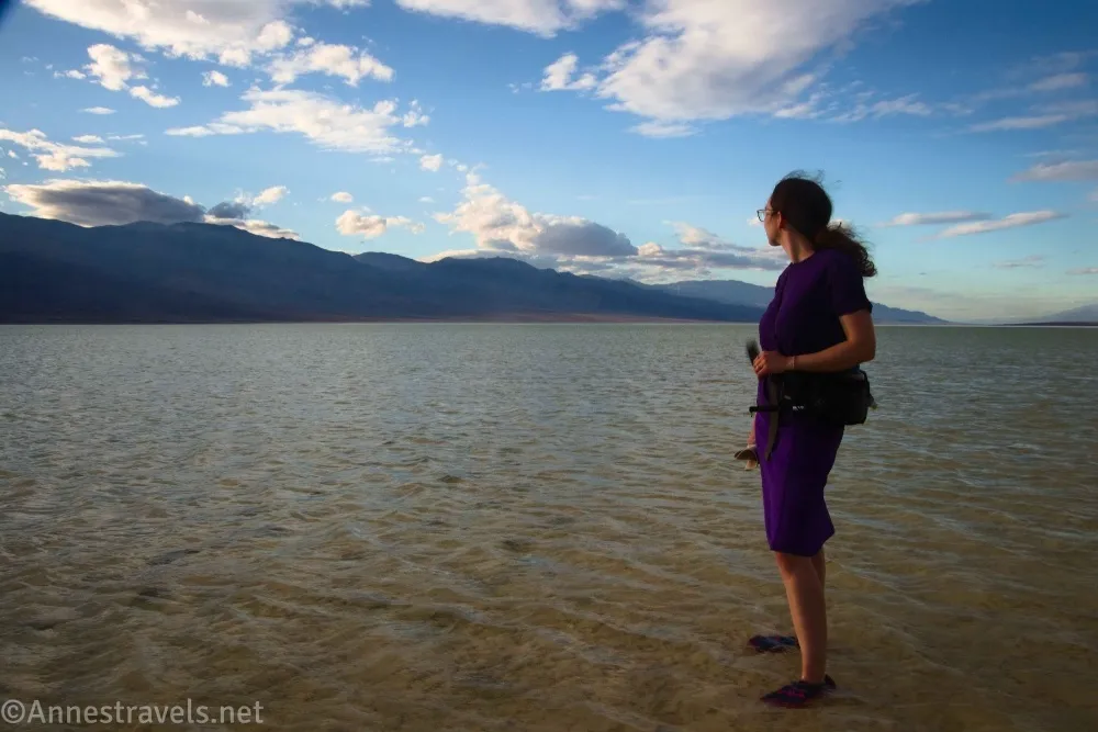 A hiker standing in a shallow lake with distant mountains and blue skies and clouds in the sunset