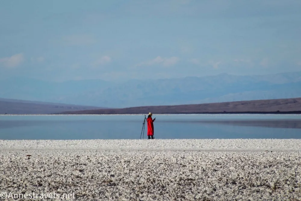 A photographer in a red jacket on a white salt plain with a lake and desert hills beyond