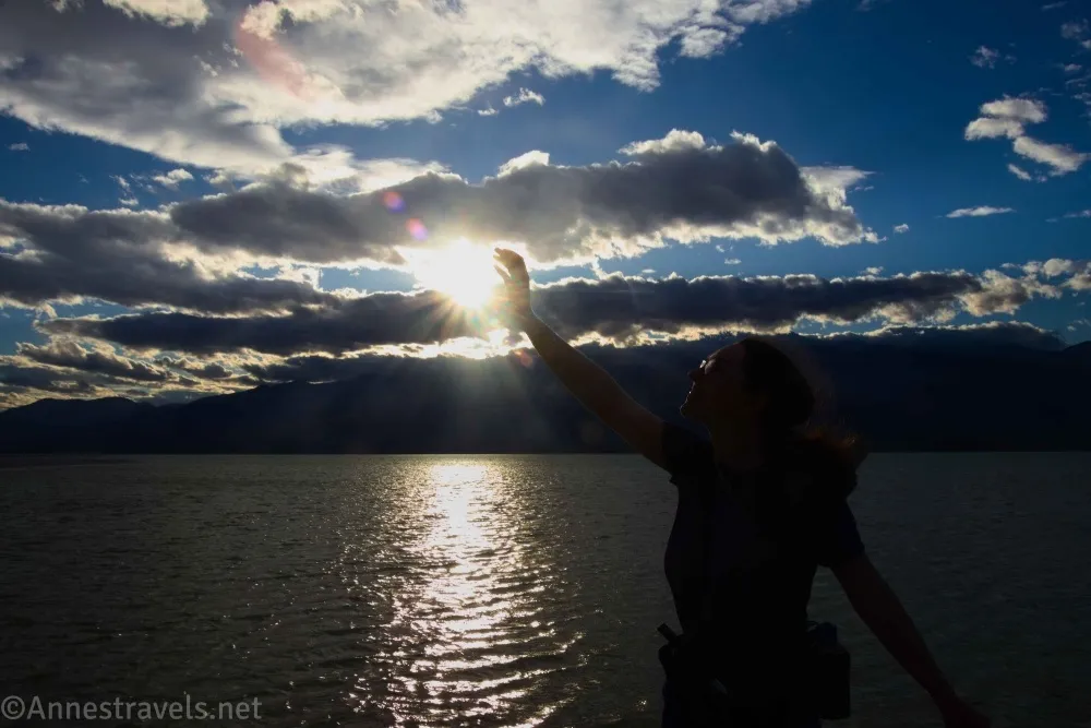 A silhouette raises a hand to the sun and clouds over a lake