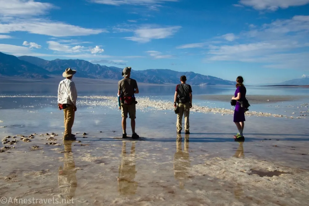 Four hikers standing in shallow water of a desert lake with distant mountains, clouds, and blue skies