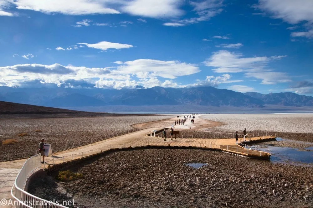 A boardwalk leading to brown and white salt flats, mountains, and a blue sky with clouds