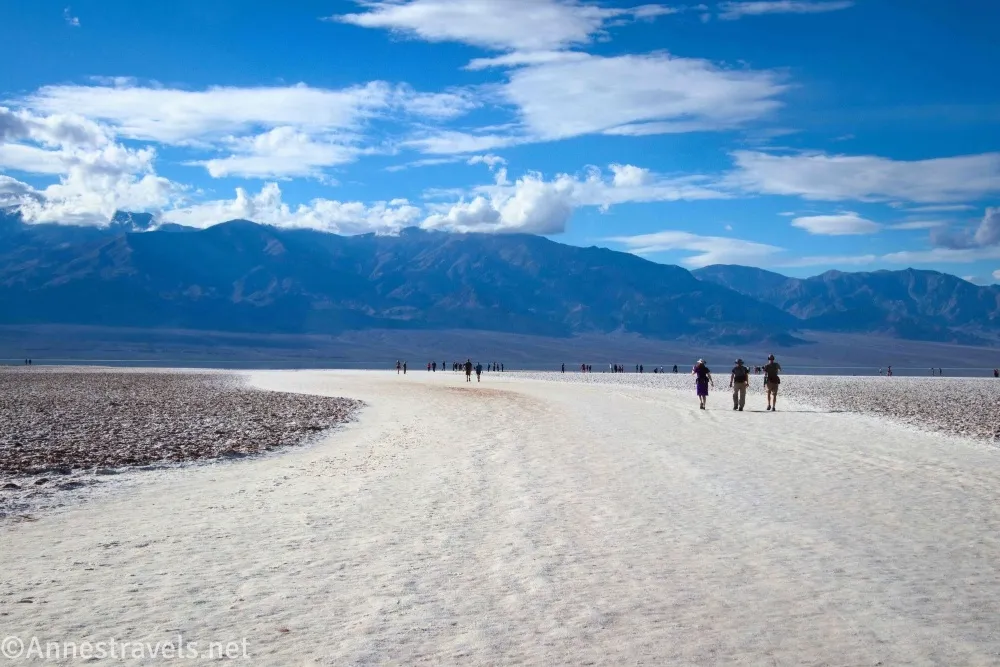 Three hikers on a white, wide path with distant mountains below blue sky and clouds