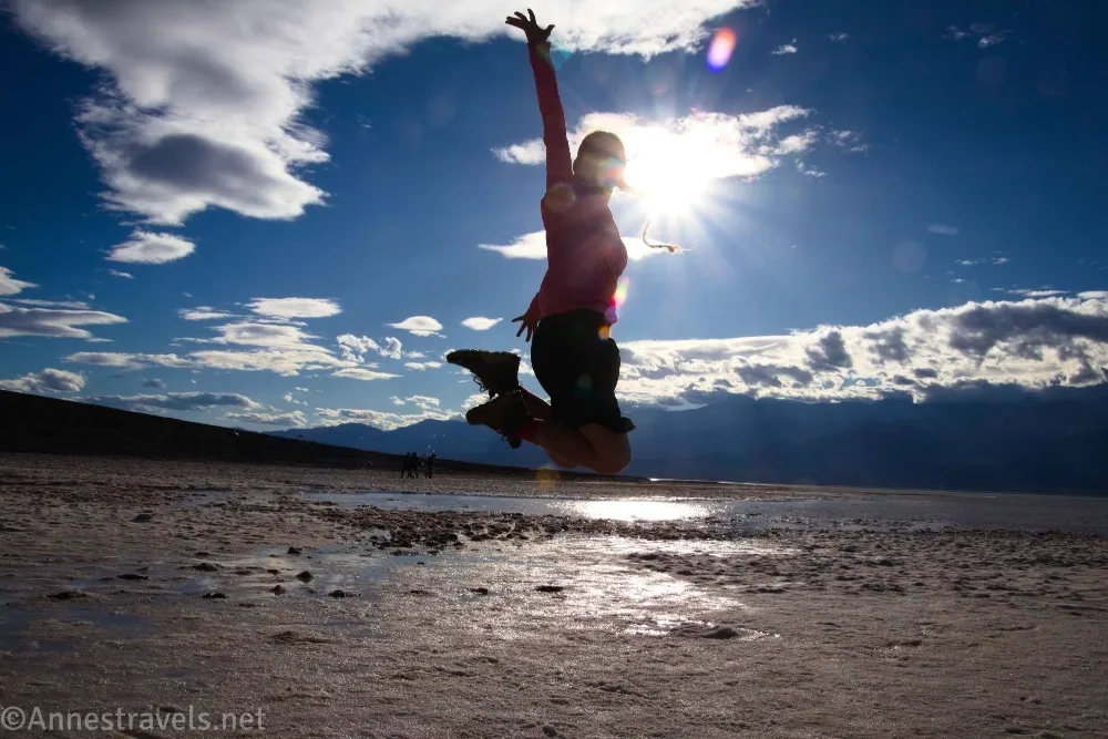 A hiker with boots and a braid jumps above a lake with the sun behind her and blue sky, clouds, and mountains in the distance