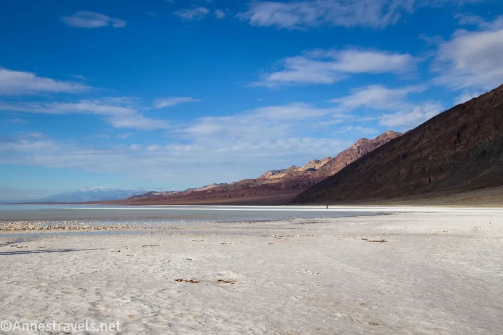 A white salt flat beach and a lake below colorful brown mountains, blue sky, and clouds