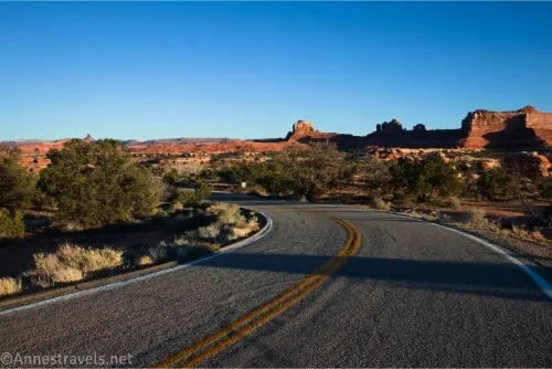 A blacktop road with two yellow center stripes with slickrock rock formations in the background