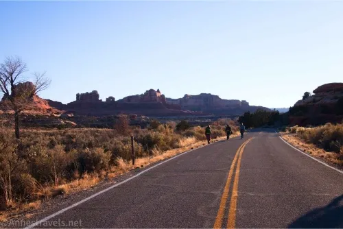 Three hikers on a blacktop road with yellow center stripes with brush and rock formations beside the road
