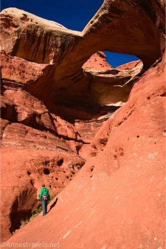 A hiker stands on a red cliff face below a giant arch