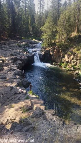 A stream falls into a pool in a waterfall between rocky cliffs and trees