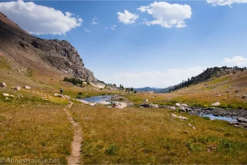 A trail meanders through golden meadows beside a blue stream with cliffs on the left and clouds in the sky 