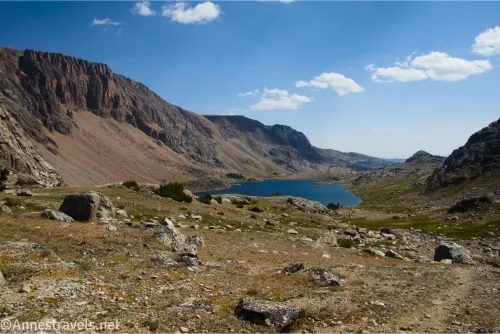 A trail in a rocky alpine meadow above a blue lake with cliffs on the left and clouds in the sky 