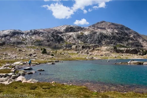 A fisherman beside a blue lake sitting among meadows in front of rocky hills 