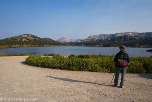 A hiker stands in a gravel parking lot beside green bushes next to a lake over which we can see mountains