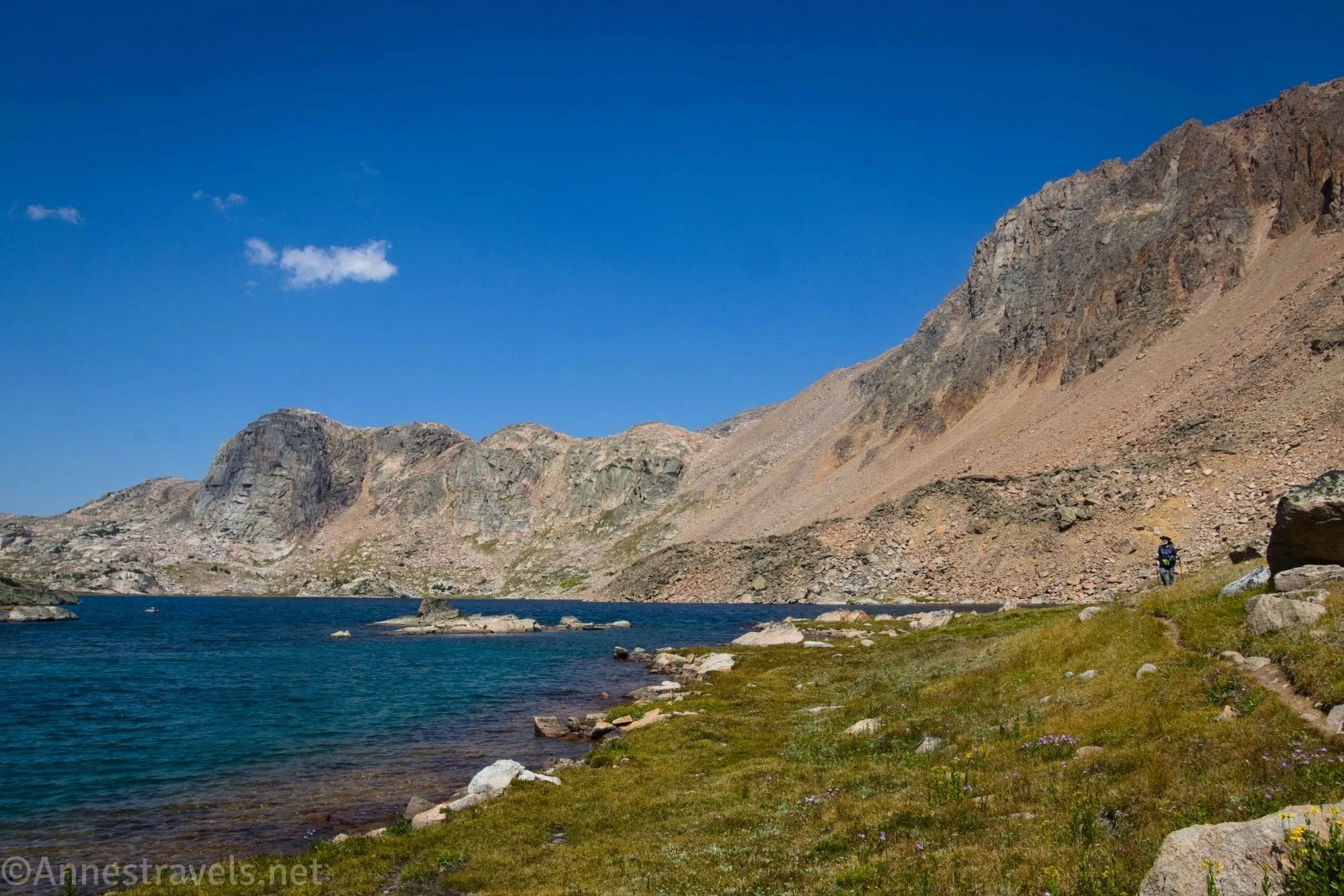 A hiker in a meadow beside an alpine lake with cliffs beyond 