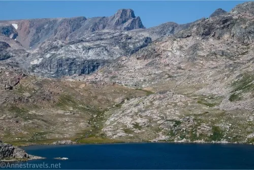 Rocky meadows and cliffs lead from a blue lake to a square peak against blue sky 