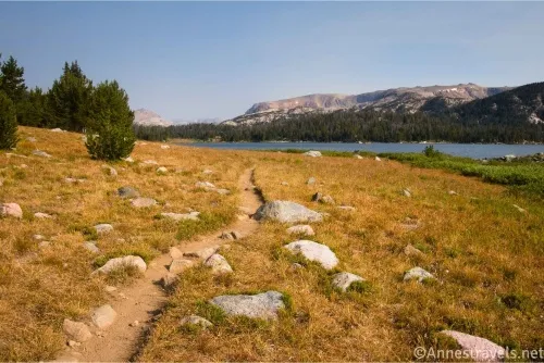 A trail through rocky meadows heading for a blue lake and distant mountains 