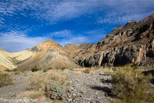 Yellow badlands and brown canyon walls above a gravelly and brushy wash and below blue skies with clouds 