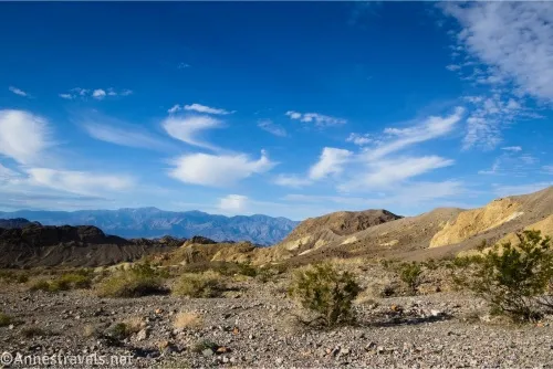 Gravelly desert and brush gives way to yellow and brown badlands and distant dark mountains below blue skies with wispy clouds