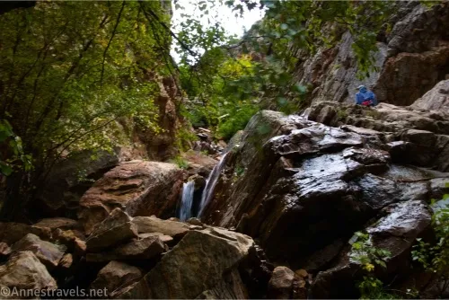A small waterfall tumbles over rocks with trees above and a person in a blue jacket in the distance 