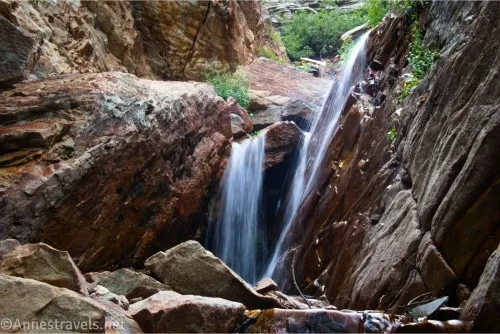 Two small waterfalls tumble over rocks with distant cliff walls and greenery 