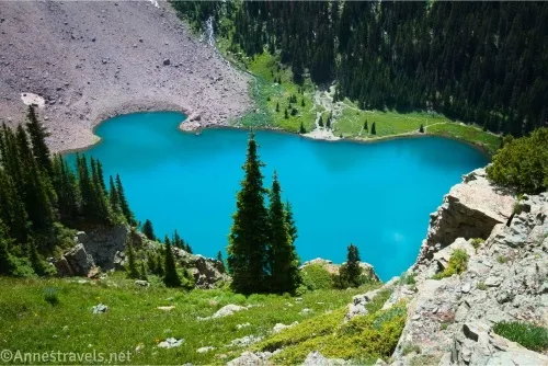 Rocks and meadows fall away toward a very blue lake with meadows, a rockslide, and forests on the shore