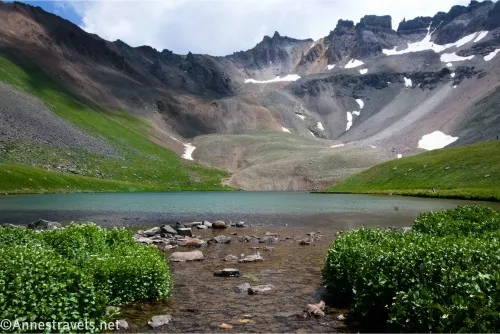 Rocky crags rise from a lake and meadows with bushes in the foreground