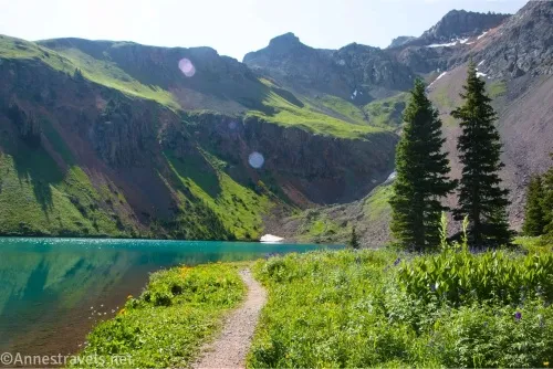 A trail in a meadow beside a lake leads past two fir trees with mountains beyond the lake