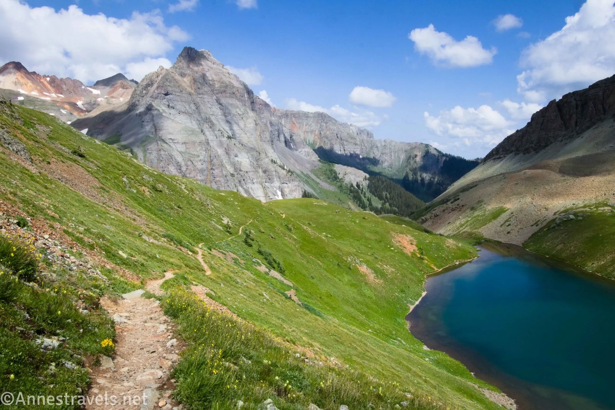 Clouds sail above mountains and a trail on a meadowed hillside above a blue alpine lake