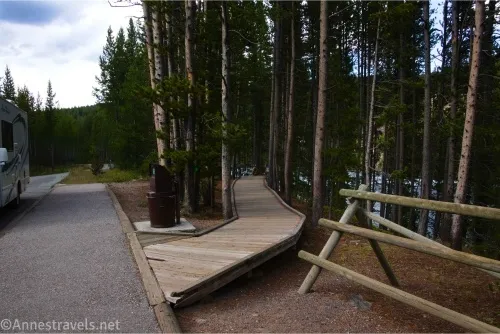 A fence and boardwalk between trees beside a sidewalk mark the trailhead