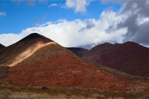 A red hill near dark hills below a blue sky with white and gray clouds