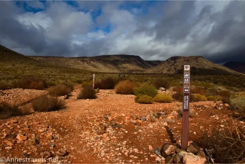 Two brown signs on either side of a rocky trail on a desert plain with distant mountains and clouds in the distance 