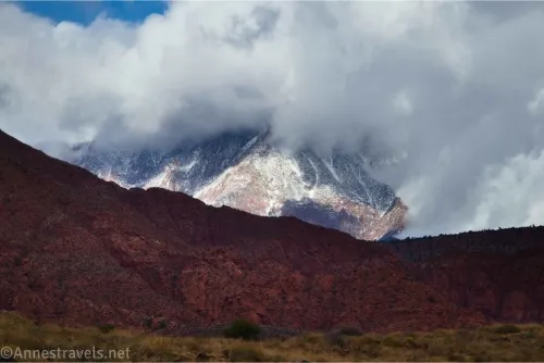 Clouds billow around a snowy mountain with dark hills in the foreground