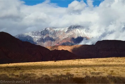 Clouds hang over mountains with fresh snow above shadow hills and a yellow, grassy plain